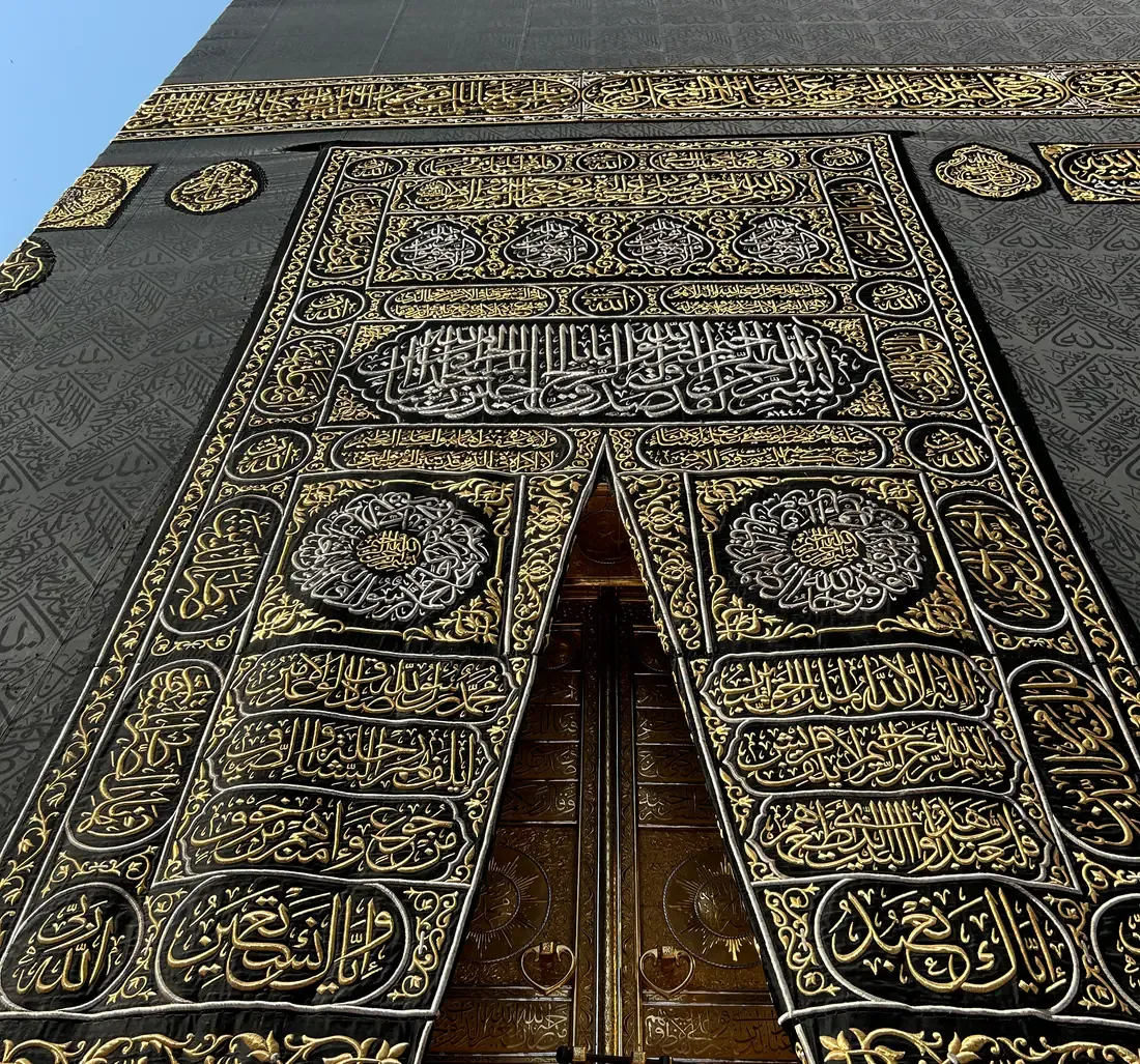 Uppåtvänd bild av en svart vägg täckt av tyg med broderad arabisk skrift i guld och silver; ett rikt broderat förhänge hänger över en förgylld dubbeldörr.