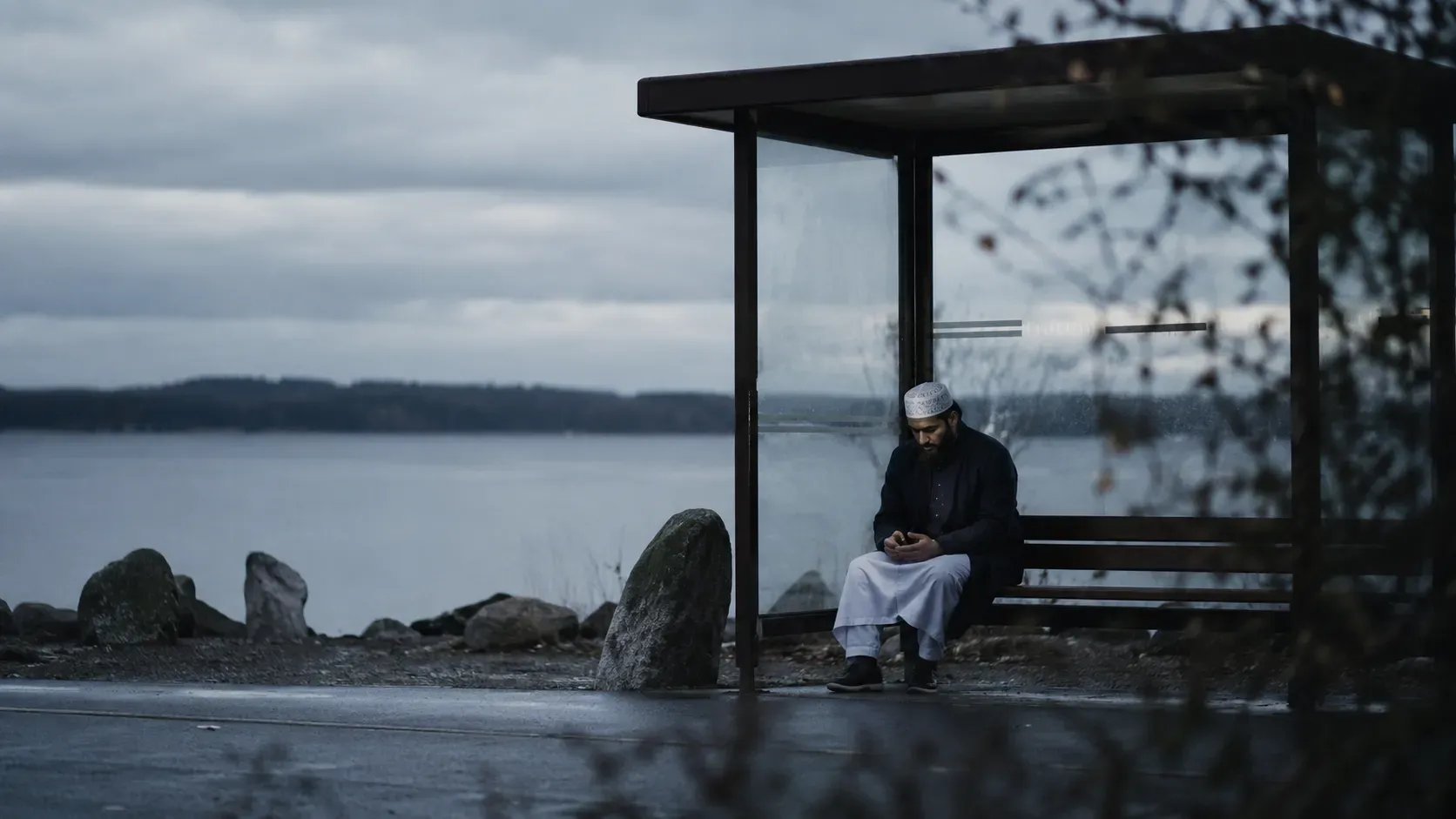 Ensam man i vit bönemössa på en busskur vid en grå svensk sjö, blicken sänkt mot telefonen.