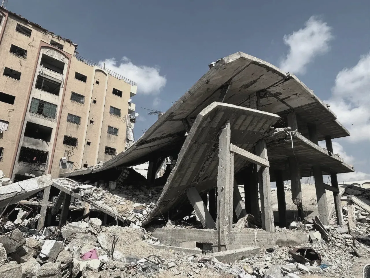 Resterna av ett bombat bostadshus i Gaza, med taket nedfällt över våningarna under och en flerfamiljsbyggnad kvar i bakgrunden.