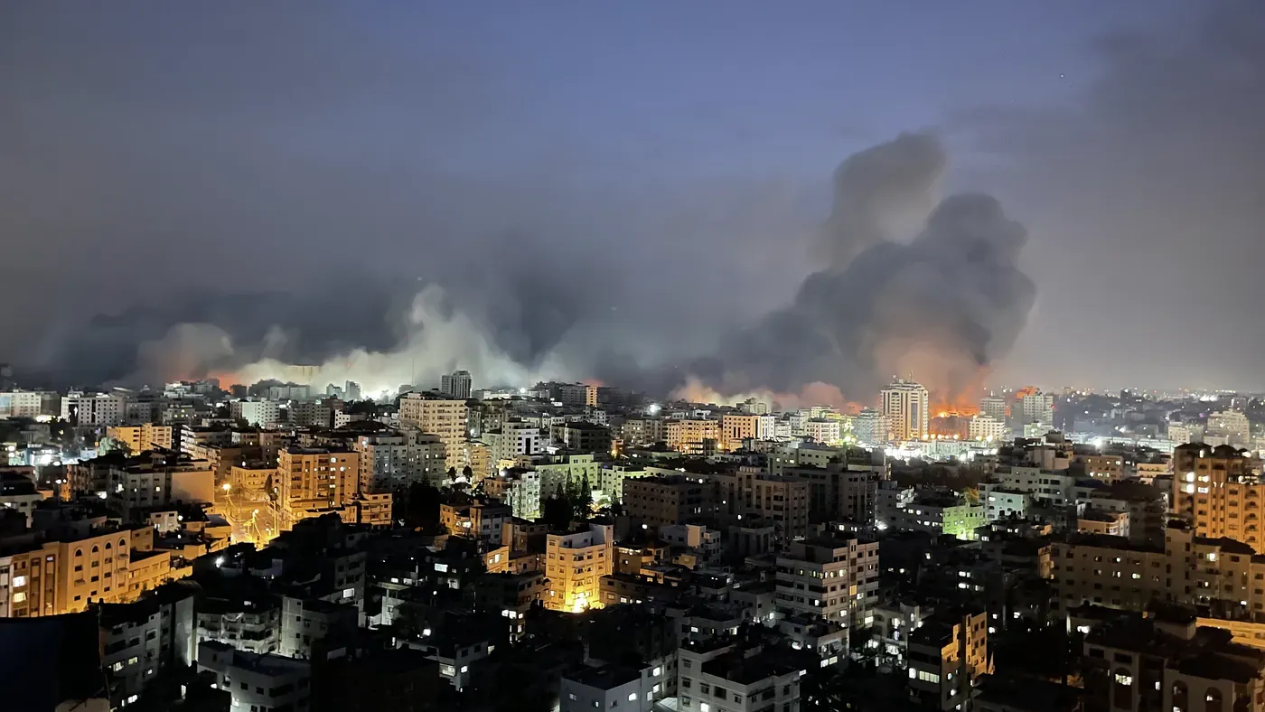 Gaza stad i skymningen sedd från ett högt hus: tända fönster och gatljus i förgrunden, och längs horisonten bränder och tjock rök efter flyganfall.