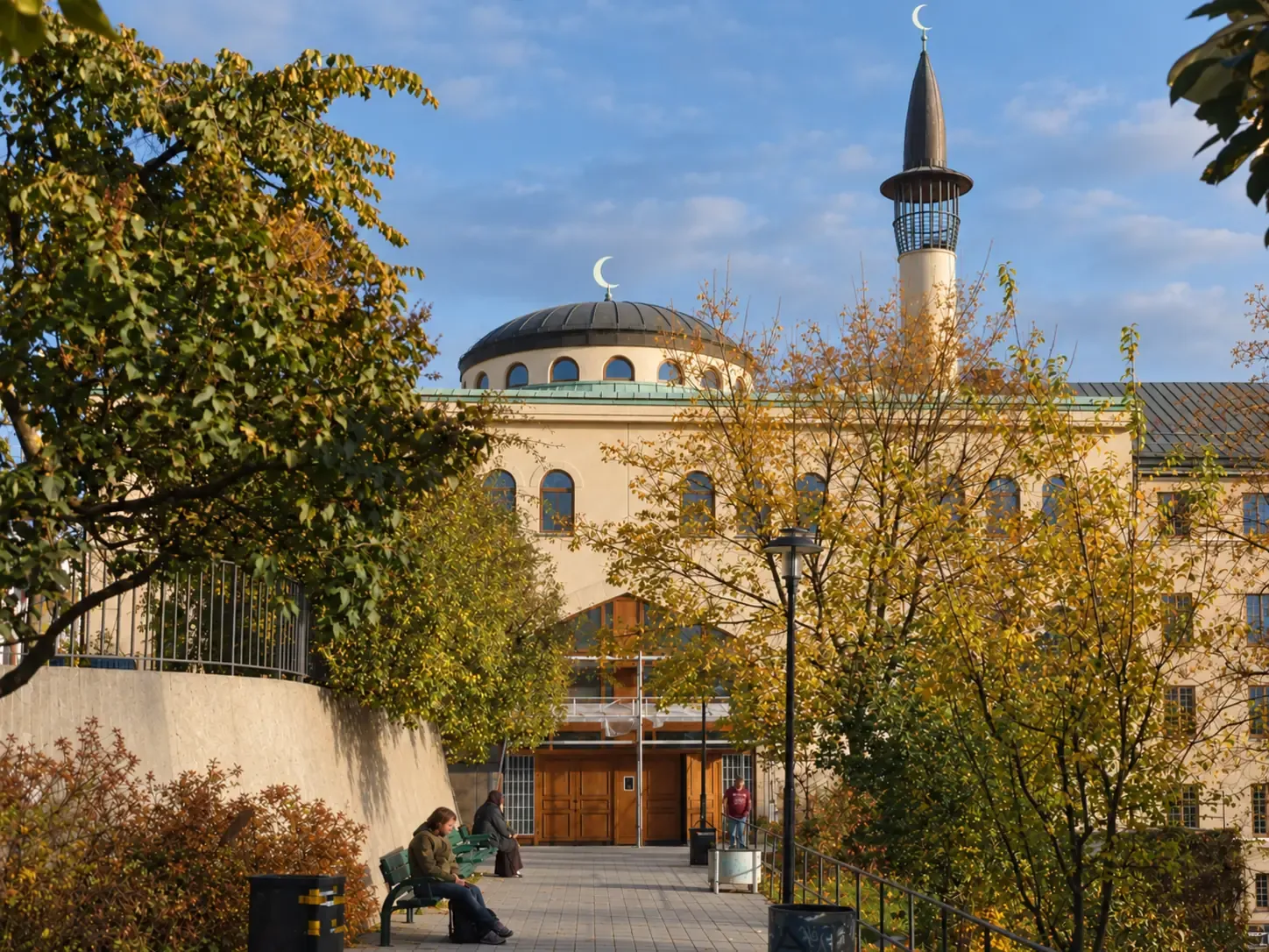 Stockholms moské på Södermalm en höstkväll, med kupolens och minaretens halvmånar mot blå himmel och gulnande lövverk framför entrén