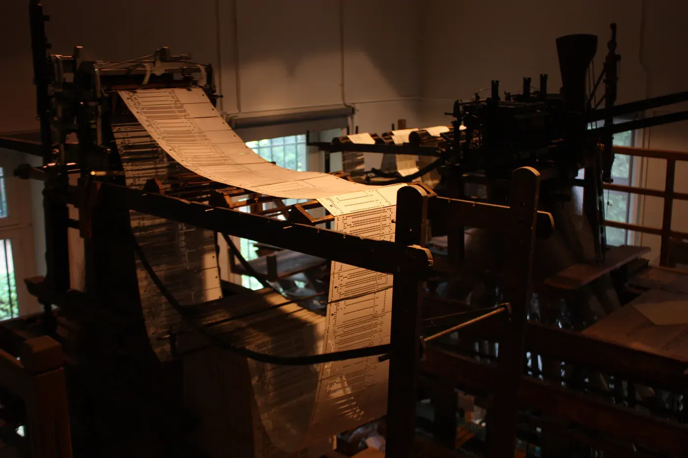 En jacquardvävstol av trä i ett halvmörkt museirum; en lång kedja av ljusa hålkort löper över maskinen i en båge och hänger ned framför den. Dagsljus faller in genom ett fönster i bakgrunden.