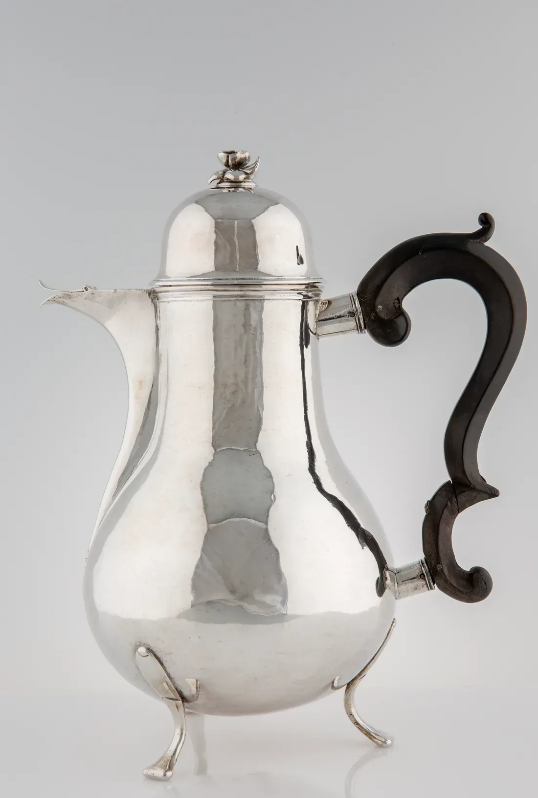 En päronformad kaffekanna i blank, slät silver på tre böjda ben, med kupigt lock med en liten blomknopp, kort pip och ett svart S-format handtag av trä.