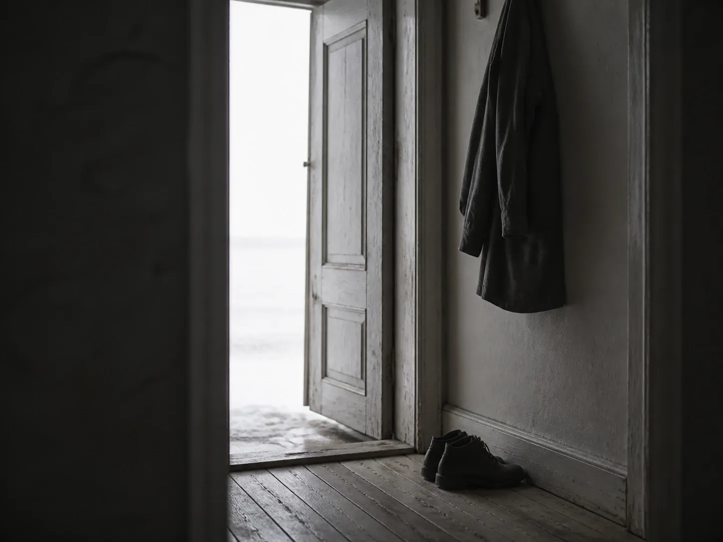 Öppen dörr från en mörk hall ut mot blekt vinterljus, med en rock på kroken och skorna avställda innanför