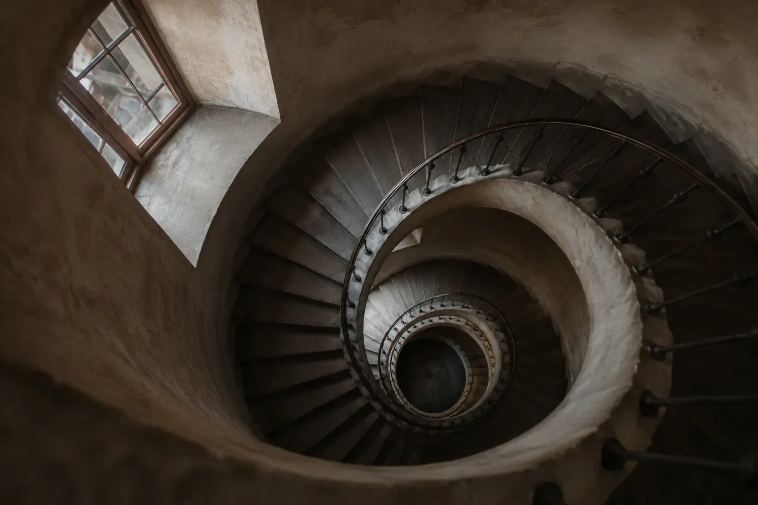 Blick rakt ner i en stenlagd spiraltrappa som vindlar nedåt i allt mindre cirklar, med ett fönster högst upp.