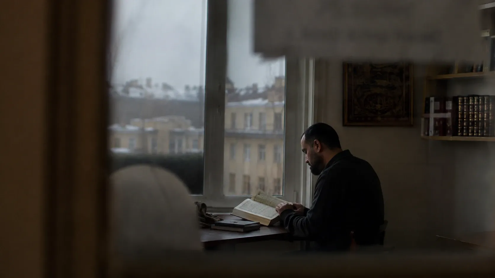 Man med kort skägg sitter vid ett skrivbord i dunkelt vinterljus och läser i en uppslagen bok, med inramad arabisk kalligrafi på väggen, inbundna band i hyllan bakom och snötäckta tak utanför fönstret.