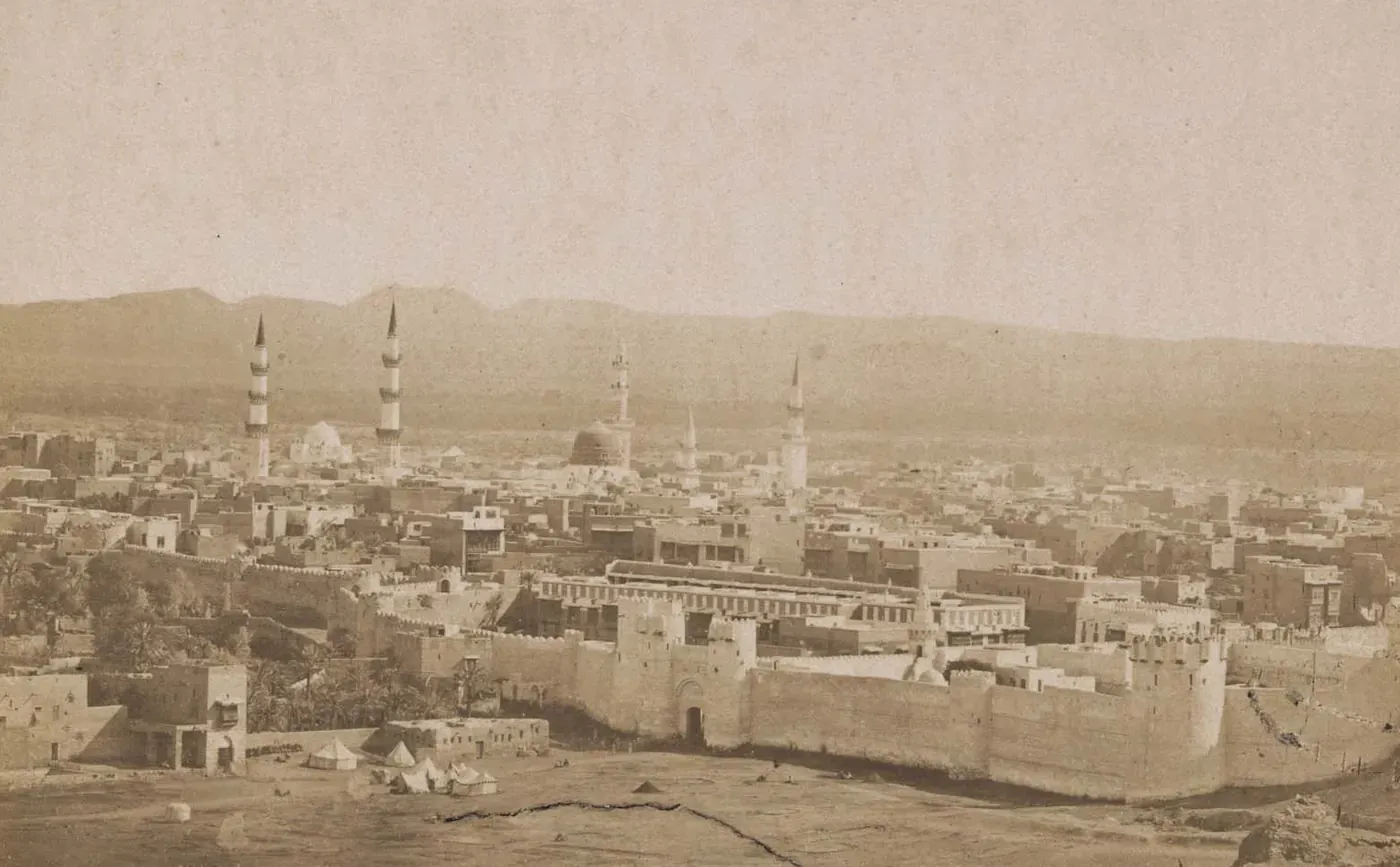 Sepiafotografi av en muromgärdad stad på en slätt, med minareter och en kupol i fjärran och berg vid horisonten.