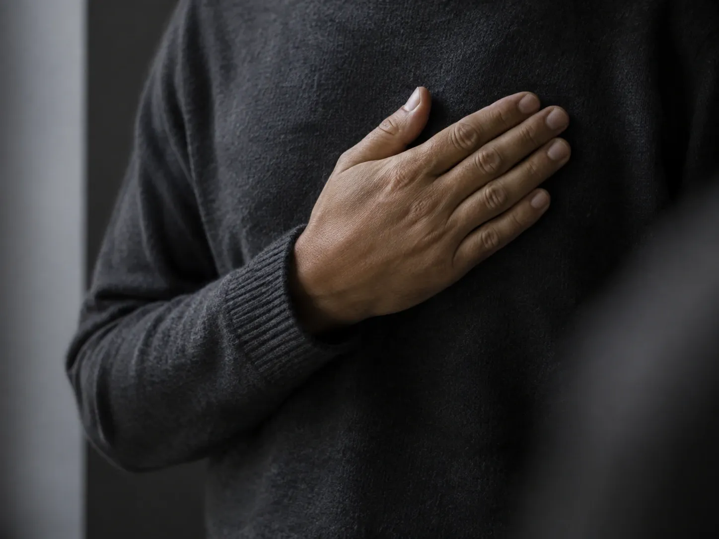 Hand lagd platt över hjärtat på en mörk ylletröja, bilden beskuren nedanför nyckelbenet