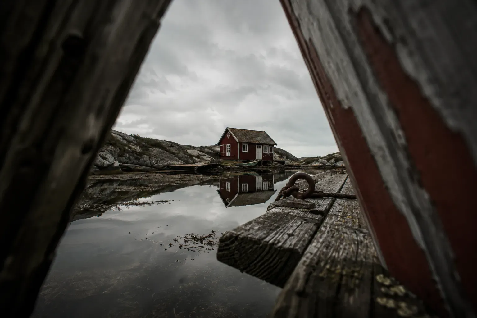 Röd skärgårdsstuga på kala klippor speglas i stilla vatten, sett genom en springa mellan vittrade träplankor.