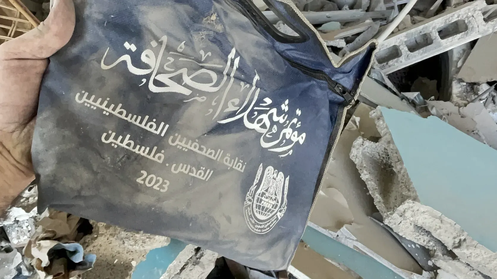 En hand håller upp en dammig blå tygväska med arabisk text i vitt, bland betongbråte, krossade väggskivor och hålsten i ett sönderbombat hus.