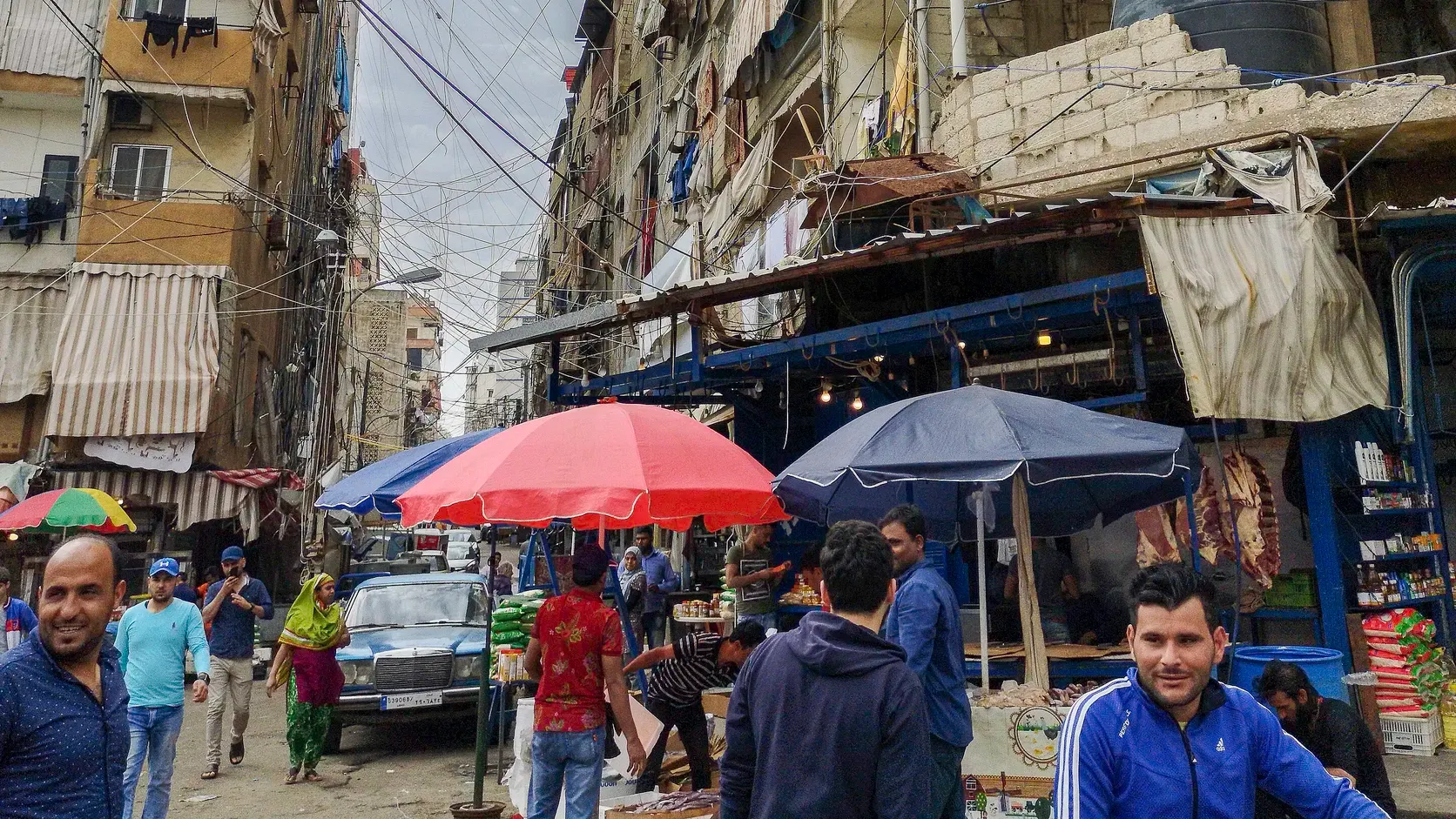 En trång marknadsgata i flyktinglägret Shatila med parasoller över stånden, slitna flervåningshus och elledningar som korsar himlen. Män går förbi, och längre bort på gatan går en kvinna i grön slöja.