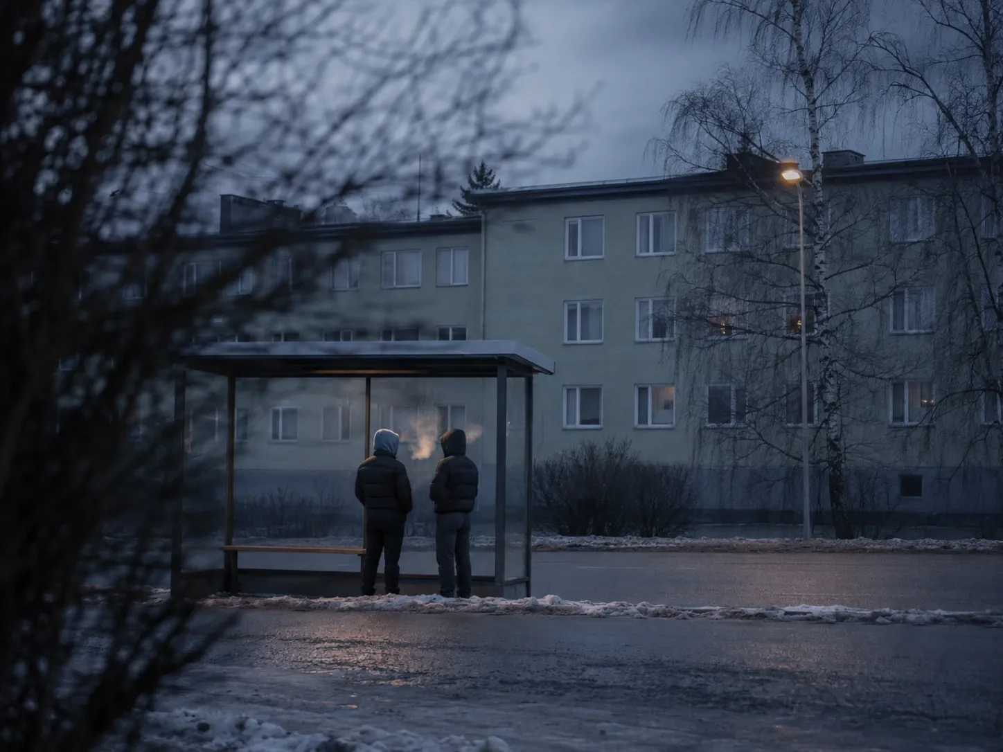 Två gestalter bakifrån vid en busskur i vinterskymning, med andedräkten synlig i kylan